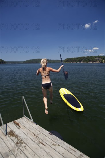 Caucasian woman jumping in lake next to paddleboard