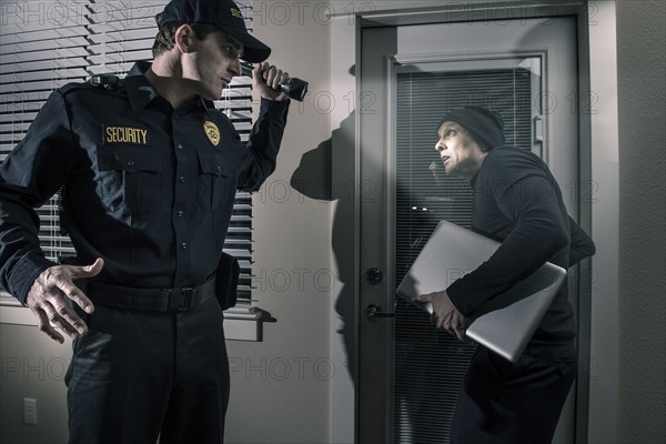 Caucasian police officer shining flashlight on burglar stealing laptop