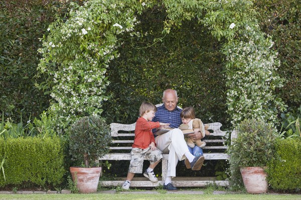 Grandfather reading book to grandchildren on garden bench