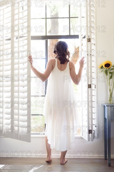 Caucasian woman wearing nightgown opening window shutters