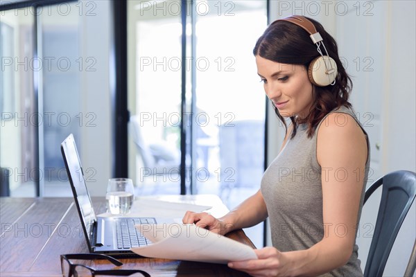 Caucasian woman listening to headphones reading paperwork using laptop