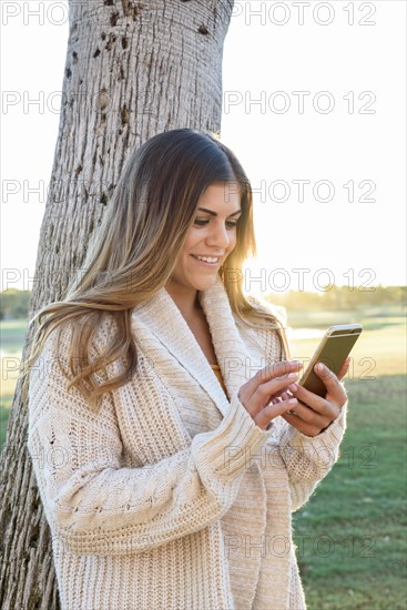 Hispanic woman leaning on tree texting on cell phone
