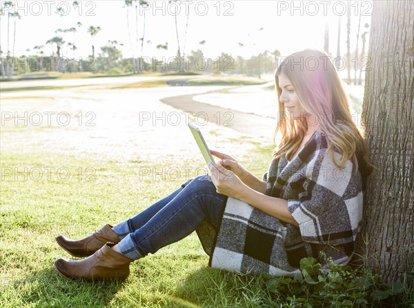 Hispanic woman leaning on tree using digital tablet