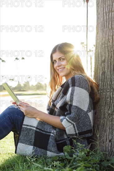 Hispanic woman leaning on tree using digital tablet