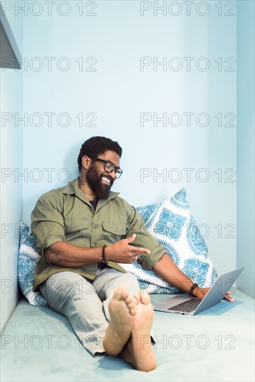 Smiling black man sitting on bed pointing at laptop