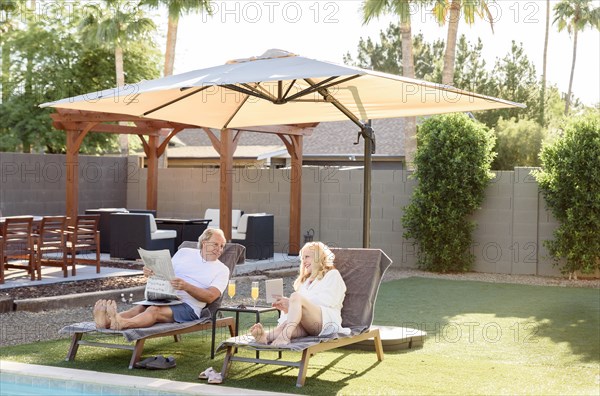 Caucasian couple reading newspaper and using laptop near swimming pool