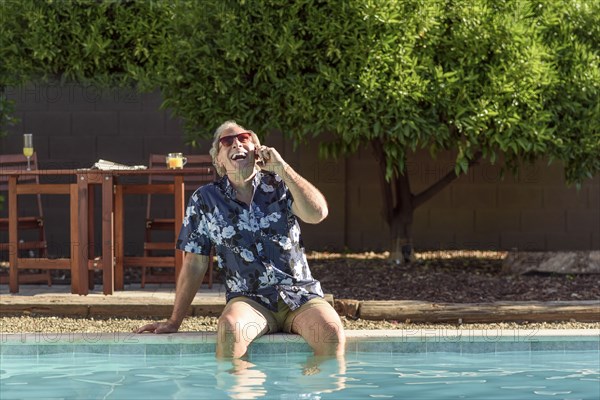 Caucasian man laughing on cell phone with legs in swimming pool