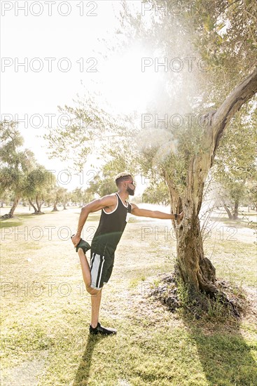 Black man leaning on tree in park stretching leg