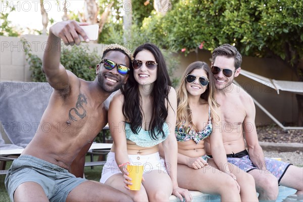 Smiling friends sitting poolside posing for cell phone selfie
