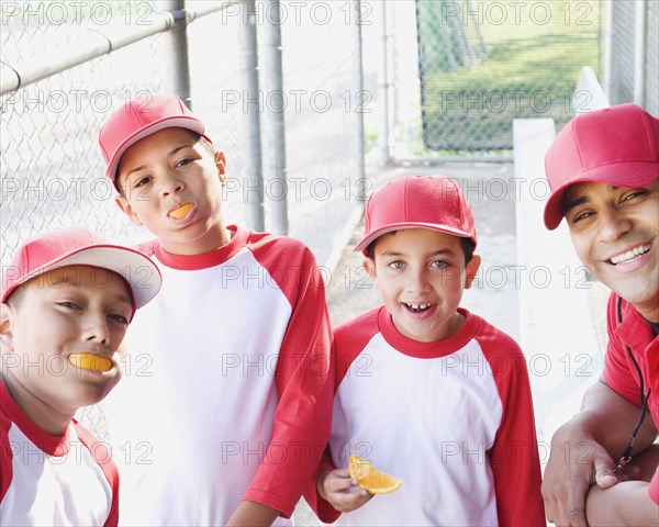 Multi-ethnic boys in baseball uniforms and coach in dugout eating oranges