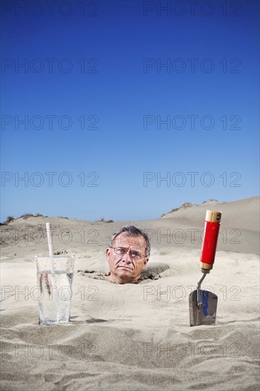 Caucasian man buried in sand next to glass of water and trowel