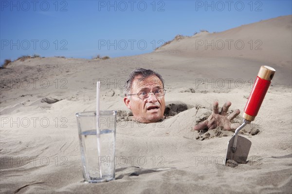 Caucasian man buried in sand next to glass of water and trowel