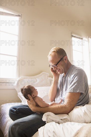 Father talking on cell phone and holding baby on bed