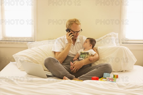 Father holding son and talking on cell phone on bed