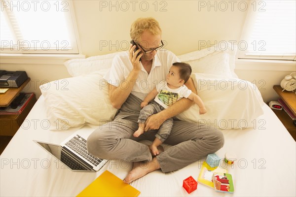 Father holding son and talking on cell phone on bed