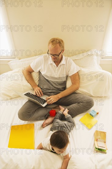 Father using laptop and playing with son on bed