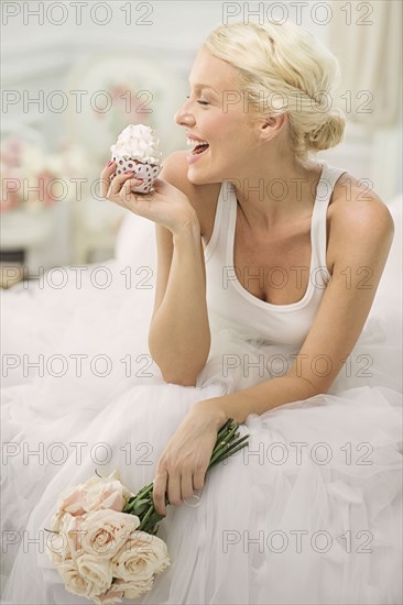 Smiling bride eating cupcake on bed