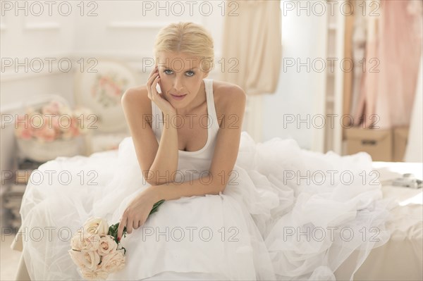 Serious bride sitting with bouquet on bed