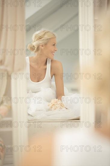 Smiling bride sitting on bed with bouquet in mirror