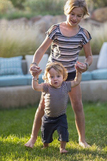 Caucasian sister helping baby brother walk in backyard