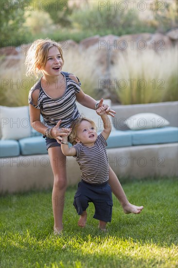 Caucasian sister helping toddler brother walk in backyard