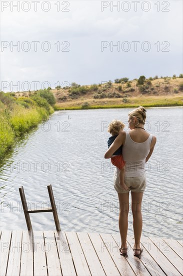 Caucasian mother holding baby son on wooden deck near lake