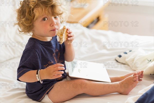 Caucasian baby boy eating and using digital tablet on bed