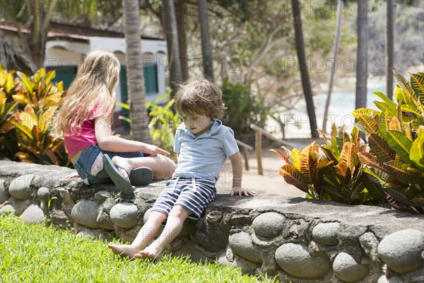 Caucasian brother and sister sitting on stone wall at beach