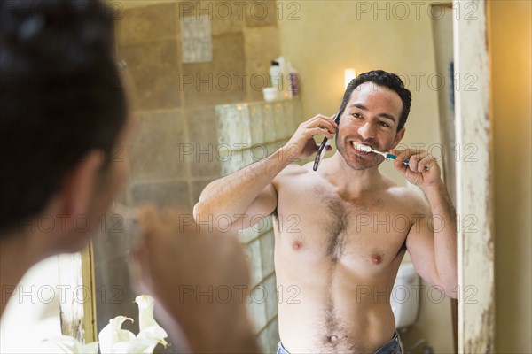 Hispanic man using cell phone and brushing teeth in mirror