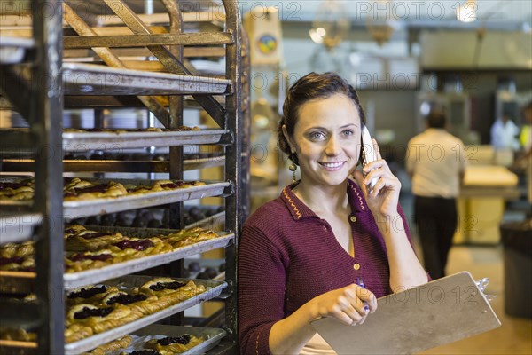 Caucasian woman holding clipboard using telephone in bakery