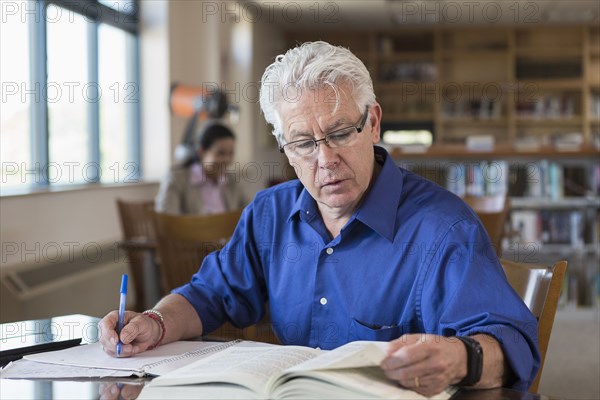 Older man reading book in library and writing notes