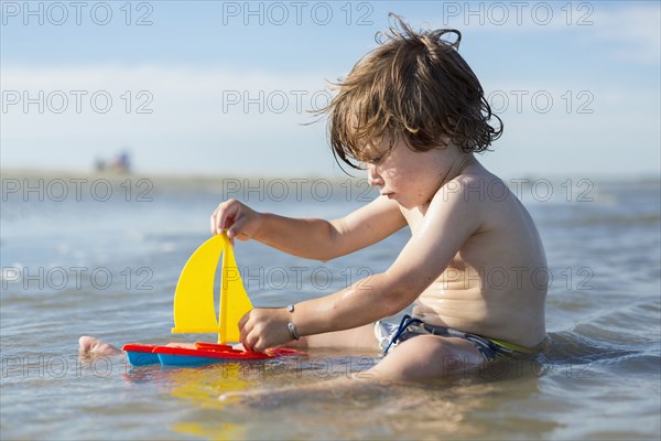 Caucasian boy sitting in ocean playing with toy sailboat