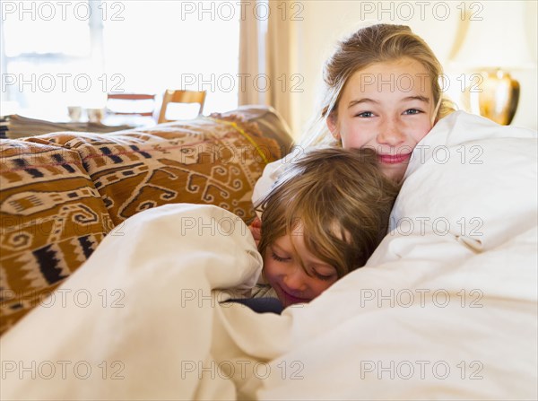 Caucasian brother and sister cuddling in comforter