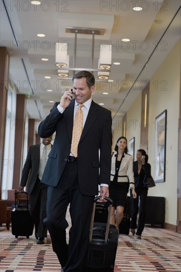 Businessman talking on cell phone and pulling suitcase down corridor