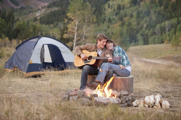 Caucasian man playing guitar for wife near campfire