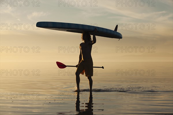 Caucasian man walking in water carrying paddle board