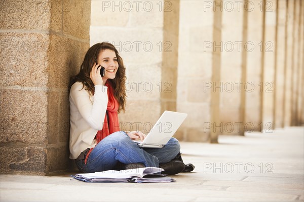 Caucasian woman sitting on ground with laptop and talking on cell phone