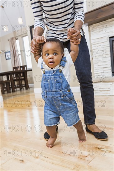 Black mother helping baby son walk on living room floor