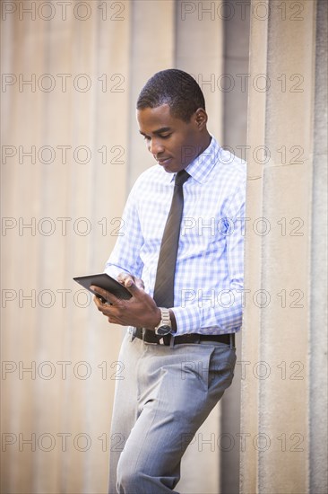 Black businessman leaning on wall using digital tablet