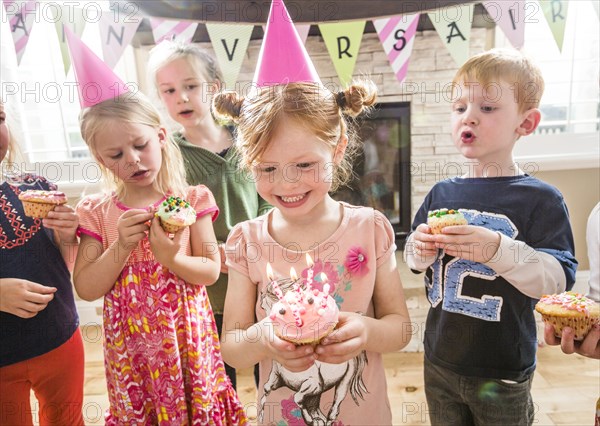 Caucasian girl wearing party hat holding cupcake with burning candles