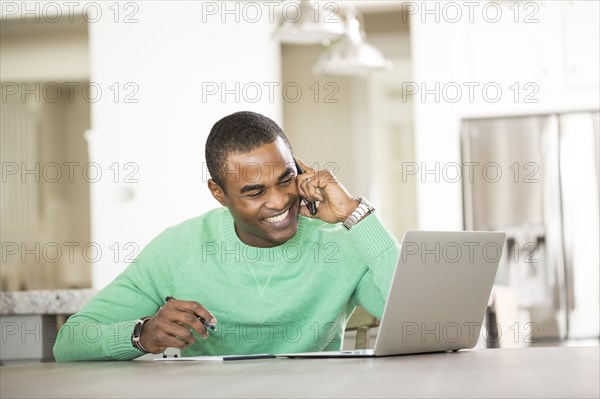 Black man sitting at table with laptop talking on cell phone