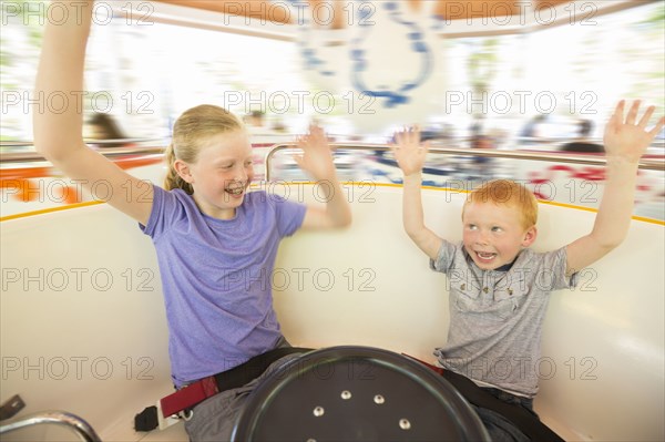 Caucasian brother and sister with arms raised on amusement park ride