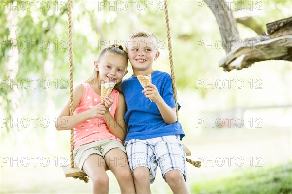 Caucasian brother and sister eating ice cream cones on rope swings