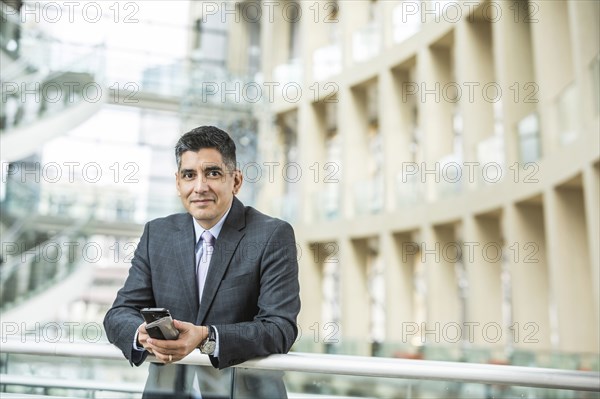 Mixed Race businessman leaning on railing in lobby holding cell phone