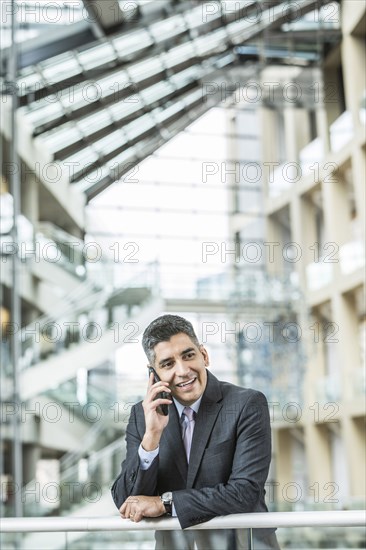 Mixed Race businessman leaning on railing in lobby talking on cell phone