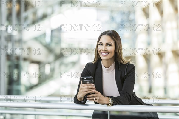 Mixed Race businesswoman texting on cell phone leaning on railing in lobby