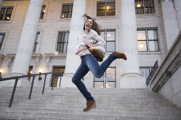 Caucasian woman carrying backpack jumping in snow on staircase