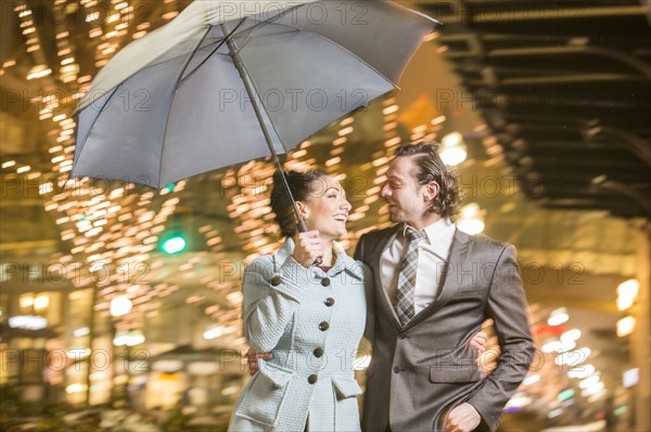 Caucasian couple carrying umbrella in city at night