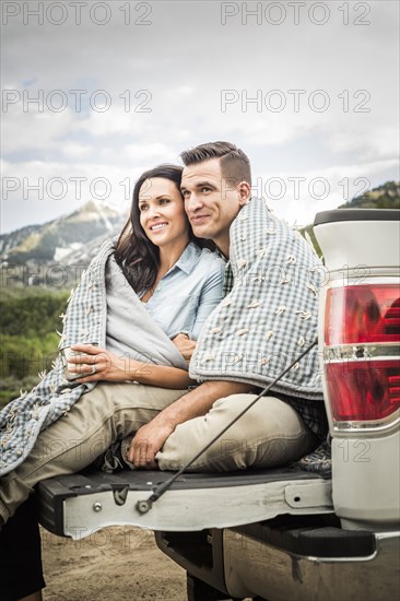 Couple wrapped in blanket sitting on bed of pick-up truck