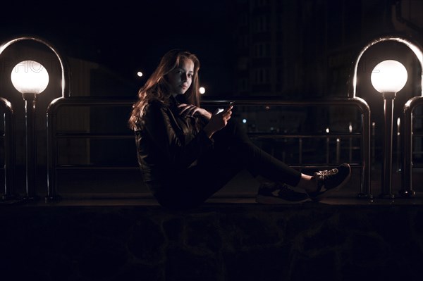 Caucasian teenage girl sitting on bench at night texting on cell phone